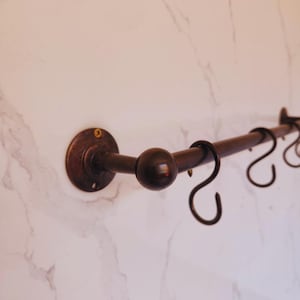 May include: A dark brown metal towel rack with a round ball in the center and two curved hooks. The rack is mounted on a white marble-patterned wall. The rack has a circular mounting plate.
