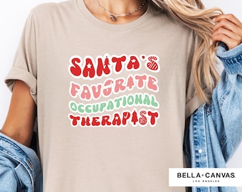 May include: A beige t-shirt with the text "Santa's Favorite Occupational Therapist" in red, pink, and green letters with a Christmas tree and star design.
