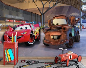 May include: A red toy car with a smiling face and the words "Lightning McQueen" on the side, next to a rusty orange toy truck with a smiling face. The toys are in front of a mural of a cityscape with a mountain in the background.