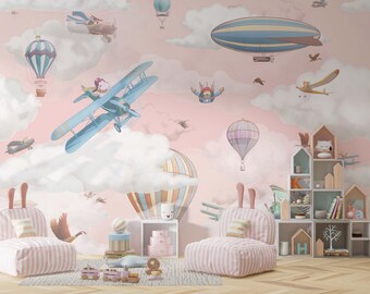 Könnte beinhalten: Ein rosa und blaues Kinderzimmer mit einer Wandtapete, die Wolken, Heißluftballons und Flugzeuge zeigt. Es gibt zwei rosa-weiß gestreifte Sessel mit Kissen und ein Holzregal mit Regalen, die mit Spielzeug und Büchern gefüllt sind.