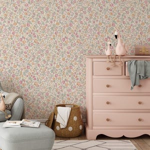 Könnte beinhalten: Ein Kinderzimmer mit floraler Tapete in Pastelltönen. Eine rosa Kommode mit goldenen Knöpfen ist mit Flamingo-Figuren geschmückt. Ein Weidenkorb mit weißen Punkten und ein grauer Stuhl vervollständigen die Szene.