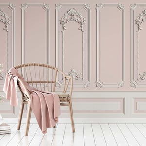 Op de afbeelding: Een lichtroze muur met witte sierlijsten en een rieten stoel met een roze deken. De stoel staat voor de muur en de vloer is wit.