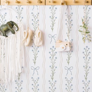 Puede incluir: Escena de pared de guardería con un atrapasueños de macramé blanco con una figura de elefante verde. Otras decoraciones incluyen zapatos de bebé color crema, una cámara de juguete de madera y vegetación. El papel pintado tiene un estampado floral.