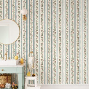 Op de afbeelding: Een badkamer met bloemenbehang met verticale strepen in crème, blauw en groen. Een ronde spiegel met een gouden frame hangt boven een mintgroene wastafel. Een witte wastafel, een gouden kraan en accessoires maken de scène compleet.