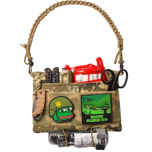 May include: A camouflage-patterned utility bag with a brown braided strap. The bag is filled with various items, including a multi-tool, a flashlight, a red first aid kit, a pair of scissors, a green frog meme patch, and a patch that says "Warzone Belgorod 2024".