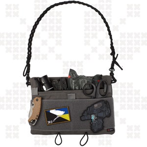 May include: A gray tactical bag with a black adjustable strap. The bag has several pockets and compartments, and is filled with various items, including a flashlight, a multi-tool, a pair of scissors, and a patch with the Ukrainian flag. The bag also has a patch with the text "WTF" on it.