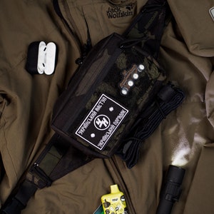Könnte beinhalten: Eine schwarze und grüne Tarn-Bauchtasche mit einem schwarzen Gurt und einem Patch, auf dem "Ukrainische Offensive" in kyrillischer Schrift steht. Die Bauchtasche befindet sich auf einer grünen Jacke mit einer schwarzen Taschenlampe und einer gelben Flasche mit einem Cartoon-Design.