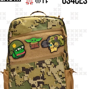 May include: A camouflage backpack with a brown zippered pocket and three patches. The patches are of a green alien, a soldier with a helmet, and a frog with a helmet. The backpack has the text "BEHEMOTH" and "US4CES" on it.