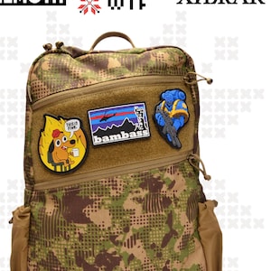 Pode incluir: Uma mochila de camuflagem verde, marrom e bege com vários bolsos com zíper e um painel de velcro para patches. O painel de patches tem três patches: um cachorro com um balão de fala que diz "This is fine", um patch azul e branco com a palavra "bambass" e um helicóptero, e um patch azul e amarelo com um robô.