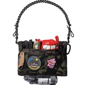 May include: A black and green camouflage utility bag with a black braided strap. The bag is filled with various items, including a red first aid kit, a multi-tool, a flashlight, and a pair of scissors. The bag has two patches, one with a skull and the text "We Are Venom", and the other with a pink skull and the text "Venom".