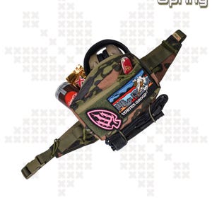 May include: A camouflage fanny pack with a variety of patches, including a pink patch with a Native American arrowhead design, a patch with the text "VORTEX GROUP", and a patch with a cityscape design. The fanny pack has a black strap and a black buckle.