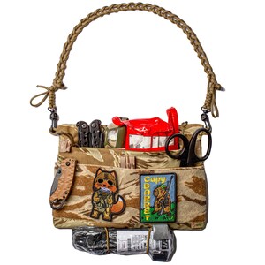 Op de afbeelding: Een bruine en beige camouflage stoffen tas met een gevlochten bruine band. De tas is gevuld met verschillende items, waaronder een rode EHBO-kit, een schaar, een multitool, een zaklamp en twee patches. Een patch toont een cartoonvos die een geweer vasthoudt en de andere patch toont een cartooncapybara die een geweer vasthoudt. De tekst "Capy Barrett" staat op de patch.