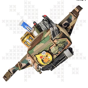 Op de afbeelding: Camouflage heuptas met meerdere vakken en compartimenten, gevuld met diverse items, waaronder een energiedrankblik, een tube blauwe gel en een multitool. Patches met tekst en afbeeldingen zijn bevestigd. De tekst "BASILISK", "WTF" en "WOODLAND M81" is zichtbaar.