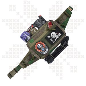 Könnte beinhalten: Eine grüne und braune Tarn-Bauchtasche mit einem Totenkopf- und Knochen-Patch, einem Zahnrad-Patch und einem roten und schwarzen Patch mit der Aufschrift "Eat the Rich". Die Bauchtasche hat einen schwarzen Gurt und einen schwarzen Reißverschluss. Die Bauchtasche befindet sich auf einem weißen Hintergrund.