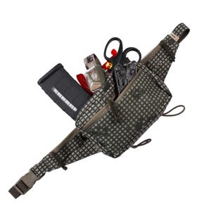 Peut inclure: Une banane de camouflage vert et noir avec une poche zippée et une poche pour chargeur noir. La banane est remplie de divers objets, dont une paire de ciseaux, un couteau suisse, une lampe de poche et un outil rouge.