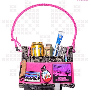 Puede incluir: Una bolsa táctica rosa y negra con una correa trenzada rosa. La bolsa está llena de varios artículos, incluyendo una lata de bebida energética Monster, una linterna, una multiherramienta y un tubo de protector solar. La bolsa tiene varios parches, incluyendo uno que dice "Warzone Belgorod 2024".
