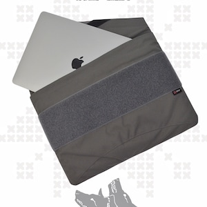 May include: A wolf grey laptop sleeve featuring a wide horizontal hook-and-loop fastener strip across the front. A silver laptop with a black apple logo is partially inserted into the sleeve. The text WTF, WOLF GREY, and CERBERUS appear.