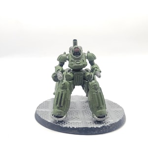 Pode incluir: Um brinquedo de robô em miniatura verde com membros articulados e um design detalhado. O robô está em pé sobre uma base circular cinza. O brinquedo é pintado em tons de verde e tem uma viseira vermelha.