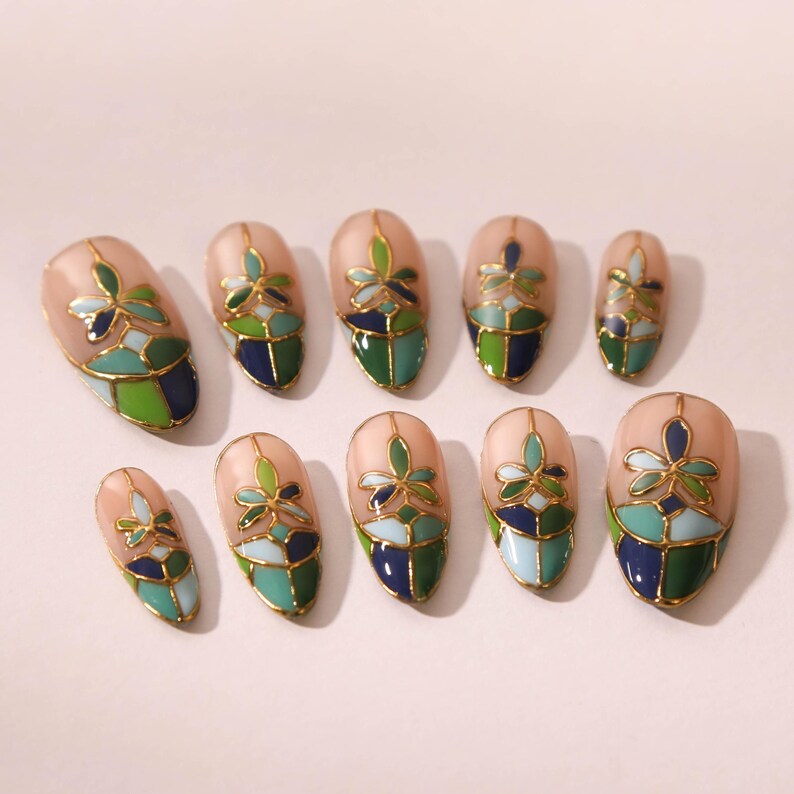 Puede incluir: Conjunto de diez u&ntilde;as postizas ovaladas con un dise&ntilde;o de vitral con detalles en verde, azul y dorado.