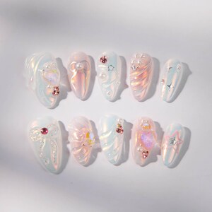 May include: A set of ten iridescent artificial nails. Each nail features a unique design with pearls, rhinestones, and star or swirl accents. The nails have a glossy finish and a light, pearlescent color palette.