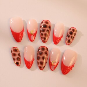 May include: Set of ten false nails with a red and brown leopard print design. Some nails have a gold crescent moon accent.
