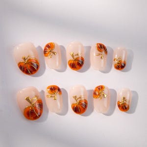 Può includere: Un set di dieci unghie finte con base rosa chiaro. Ogni unghia è decorata con un design dettagliato di zucca arancione e marrone con steli e accenti dorati. Le unghie sono di forma ovale.