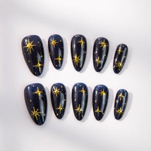 May include: A set of ten artificial nails in various sizes. Each nail is a deep navy blue with a glossy finish and adorned with gold star embellishments. The stars vary in size and placement, creating a celestial design.