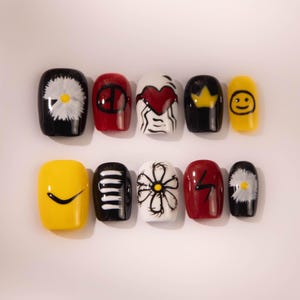 Puede incluir: Un conjunto de diez uñas postizas con varios diseños. Las uñas presentan una mezcla de colores, incluyendo negro, rojo, blanco y amarillo. Los diseños incluyen una margarita, un símbolo de la paz, un corazón, una corona, una carita sonriente y un rayo.