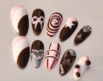 May include: Set of ten false nails with different designs. The nails are brown with pink and white accents. Some nails have a bow, a heart, a star, a butterfly, and stripes.