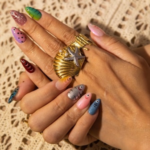May include: A hand with long, colorful, painted fingernails. The nails feature various designs, including fish, polka dots, and glitter. A gold ring with a seashell and starfish design is also visible.