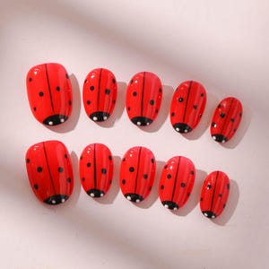 May include: A set of ten red press-on nails, each decorated to resemble a ladybug. The nails have a glossy finish with black spots and a black and white base, creating a playful and vibrant aesthetic.