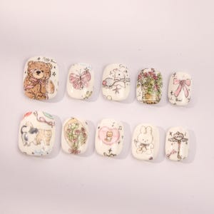 May include: Ten white square-shaped false nails with different cute cartoon designs. The designs include a teddy bear, a butterfly, a bunny, a cat, a flower bouquet, a key, and a bow.