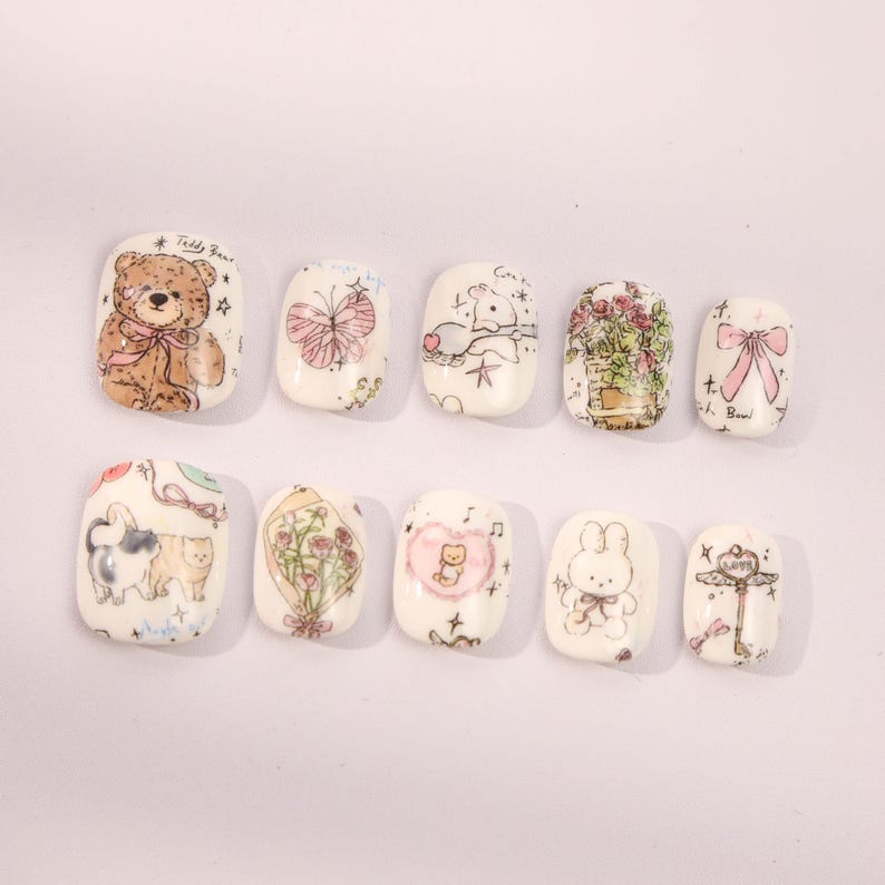 May include: Ten white square-shaped false nails with different cute cartoon designs. The designs include a teddy bear, a butterfly, a bunny, a cat, a flower bouquet, a key, and a bow.