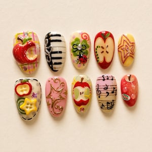 May include: A set of ten press-on nails featuring diverse designs including a red apple on a plaid background, piano keys, a ladybug on a clover, a sliced apple, a yellow star, apple-shaped buttons, gold swirls, musical notes, and red fruit.