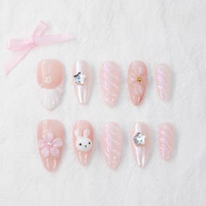 Könnte beinhalten: Set aus zehn falschen Nägeln in Rosa und Weiß mit verschiedenen Designs, darunter Blumen, ein Kaninchen und Sterne.