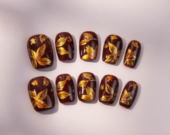 May include: Set of ten artificial nails in a dark burgundy color, each adorned with a gold leaf design. The leaves have intricate details and are accented with small silver glitter. The nails are oval-shaped.
