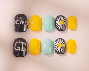 May include: A collection of ten artificial nails in different designs. The nails are in a variety of colours, including yellow, light blue, black, and white. Some nails have the word "POWER" or "GD" printed on them. Other nails have a daisy design.