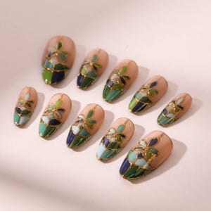 Puede incluir: Conjunto de diez u&ntilde;as postizas con un dise&ntilde;o floral. Las u&ntilde;as son de color rosa claro con un contorno dorado y detalles florales verdes, azules y blancos.