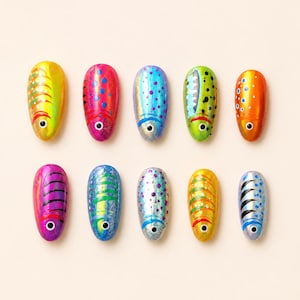 May include: Ten oval-shaped nail art press-on tips arranged in two rows of five. Each tip features a unique fish-inspired design with vibrant colors including yellow, pink, blue, green, and orange, accented with black eyes and patterns.