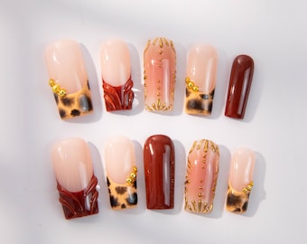 May include: A collection of artificial nails in various designs. Some nails feature a nude base with a leopard print tip, while others have a deep red French tip. Several nails are adorned with gold embellishments and a gradient pink hue.