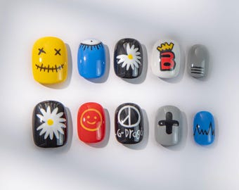 Puede incluir: Un conjunto de diez uñas postizas con varios diseños. Las uñas presentan una cara amarilla, una uña azul con un diseño de ojo blanco, una uña negra con una margarita blanca, una uña blanca con una B roja y una uña gris con rayas negras. Otras uñas incluyen una margarita negra, una carita sonriente roja y una uña negra con el texto "G-Drago".