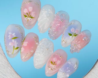 Könnte beinhalten: Ein Set mit zehn ovalen künstlichen Nägeln mit einer transparenten Basis und bemalten Blumenmustern. Die Designs beinhalten rosa, weiße und blaue Blumen mit goldenen Akzenten.