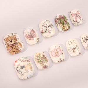 May include: Set of ten white press-on nails with various cute cartoon designs. The designs include a teddy bear, a butterfly, a bunny, a cat, a bouquet of flowers, and a heart with a cat inside.
