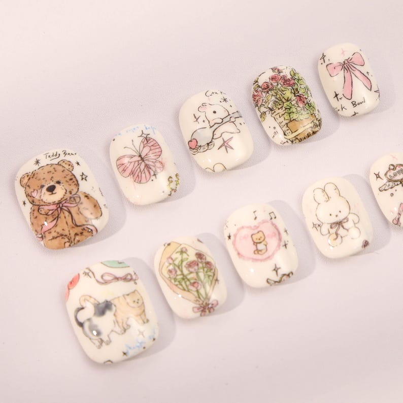 May include: Set of ten white press-on nails with various cute cartoon designs. The designs include a teddy bear, a butterfly, a bunny, a cat, a bouquet of flowers, and a heart with a cat inside.