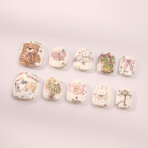 May include: Set of ten white square-shaped false nails with various cute cartoon designs, including a teddy bear, a butterfly, a cat, a bunny, a flower bouquet, and a musical note.