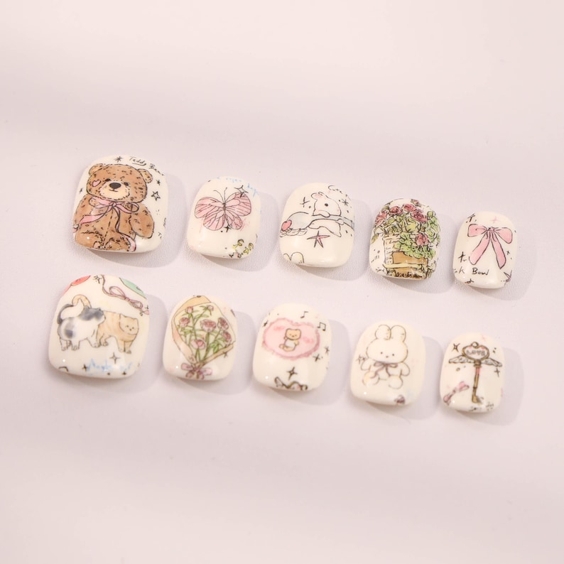 May include: Set of ten white square-shaped false nails with various cute cartoon designs, including a teddy bear, a butterfly, a cat, a bunny, a flower bouquet, and a musical note.
