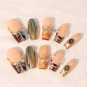 May include: Set of ten false nails with various designs. Some nails feature a gold swirl pattern, others have a brown and gold cross, and some have a brown and gold fleur-de-lis. Some nails are decorated with pearls and rhinestones.