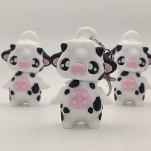 May include: Three white cow keychains with black spots and pink accents. Each keychain has a white head with black ears, large black eyes, and a pink nose. The cow's body is white with black spots and a pink udder.