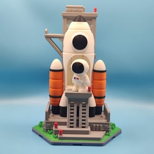May include: A white and orange toy rocket ship with a small astronaut figure standing on a launch pad. The rocket has black accents and is set against a light blue background. The launch pad is gray and green.