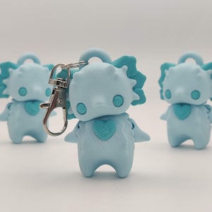 May include: Three light blue keychains shaped like a cartoon creature with a heart on its chest. The keychains have a loop at the top and a silver clasp. The creature has a spiky head and small arms.